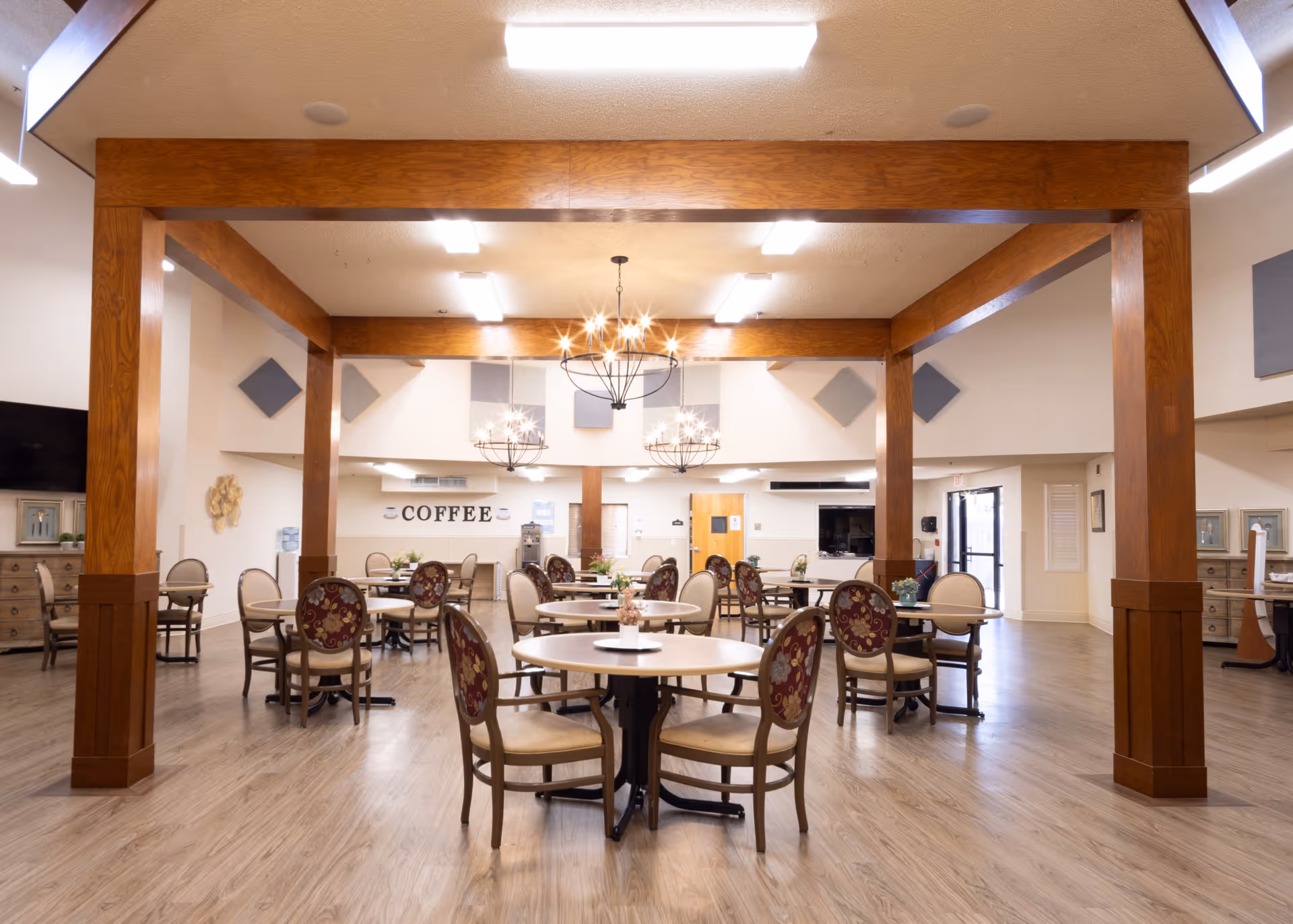A spacious dining area with multiple round tables and chairs arranged neatly. The room features wooden beams, modern chandeliers, and a sign on the back wall that reads 'COFFEE'. The floor is light wood, and there are decorative elements on the walls and furniture along the sides.