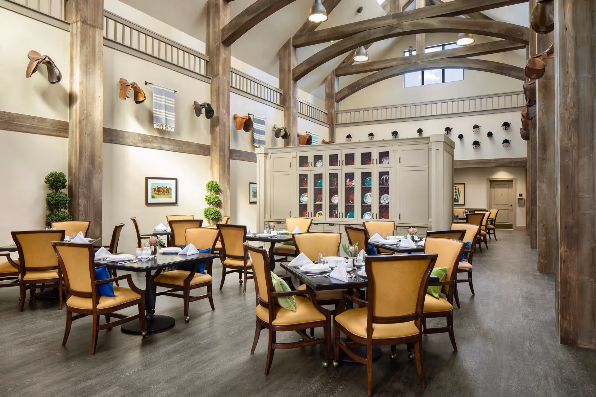 A spacious dining room with high vaulted ceilings and exposed wooden beams. The room features multiple tables set with white napkins, glassware, and small floral centerpieces. The chairs have wooden frames with tan upholstery. The walls are decorated with horse saddles, framed pictures, and small topiary plants. A large cabinet with glass doors displaying plates is positioned in the center background.