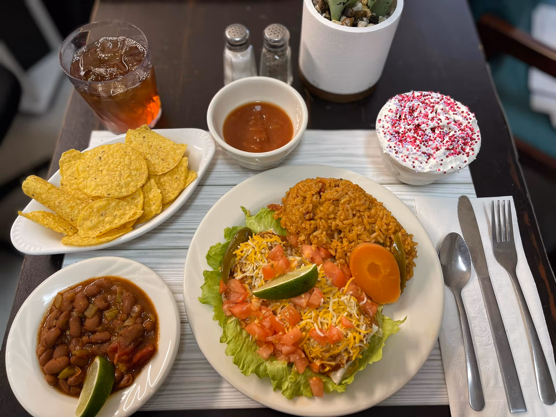 A meal served on a table including a plate with rice, a taco salad topped with shredded cheese, diced tomatoes, a lime wedge, a carrot slice, and lettuce. There is a bowl of pinto beans with a lime wedge, a bowl of salsa, a plate of tortilla chips, a glass of iced tea, and a dessert cup topped with whipped cream and red and purple sprinkles. Silverware including a fork, knife, and spoon is placed on a napkin to the right of the plate.