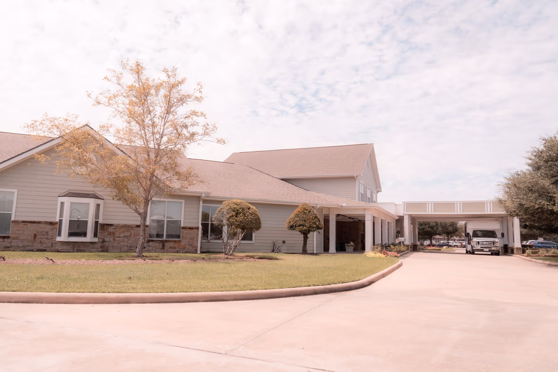 Exterior view of The Reserve at Katy Assisted Living and Memory Care building with a driveway, manicured lawn, trees, and a covered entrance where a white vehicle is parked.