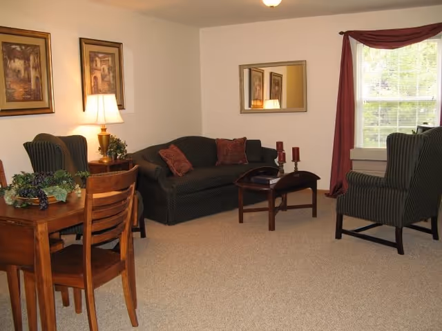 A cozy living room with a dark green sofa adorned with red pillows, two matching armchairs, a wooden coffee table with decorative candles, a wooden dining table with chairs, a table lamp, framed artwork on the walls, a large mirror, and a window with red curtains letting in natural light.