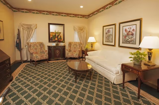 A cozy living room with a white sofa, two floral patterned armchairs, a wooden coffee table, and side tables with lamps. The walls are decorated with framed floral artwork and a mirror above a small cabinet with a flower arrangement. The room has a patterned carpet and soft lighting.