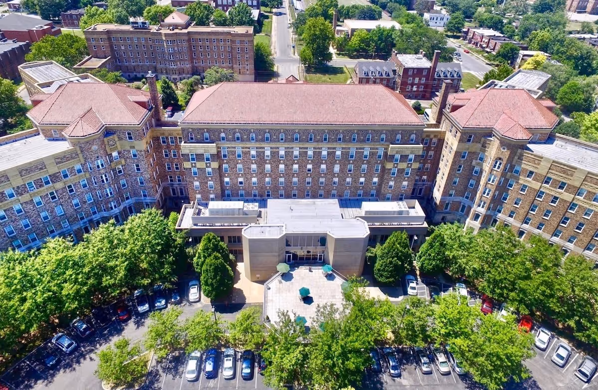 Aerial view of Homer G Phillips Senior Apartments, a large multi-story brick building with a red-tiled roof, surrounded by trees and a parking lot with several cars. The building has a central entrance area with outdoor seating and umbrellas.