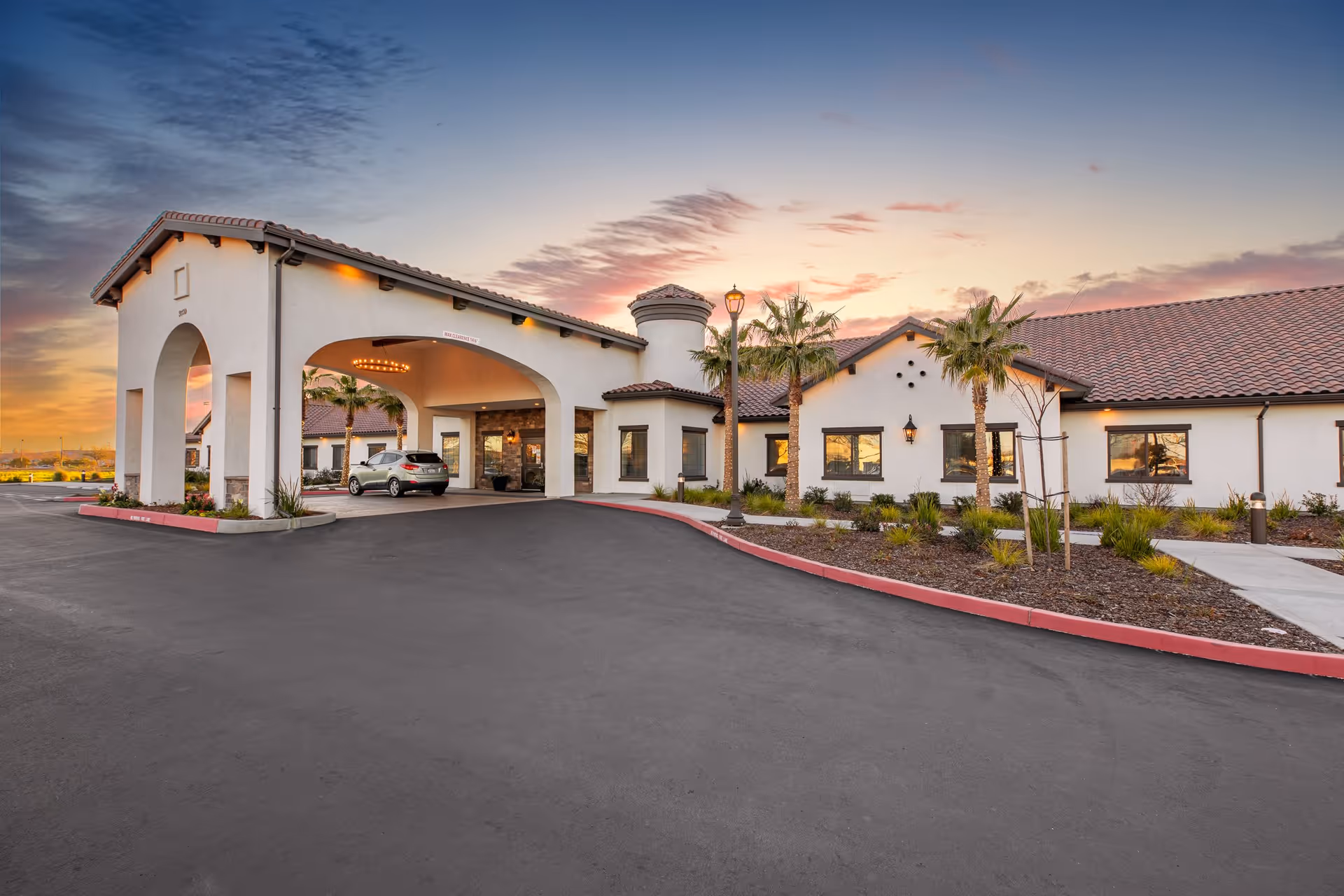 Exterior view of The Gardens At Laguna Springs facility at sunset, showing a large covered entrance with arches, a car parked underneath, palm trees, and a well-maintained landscape with shrubs and a sidewalk.