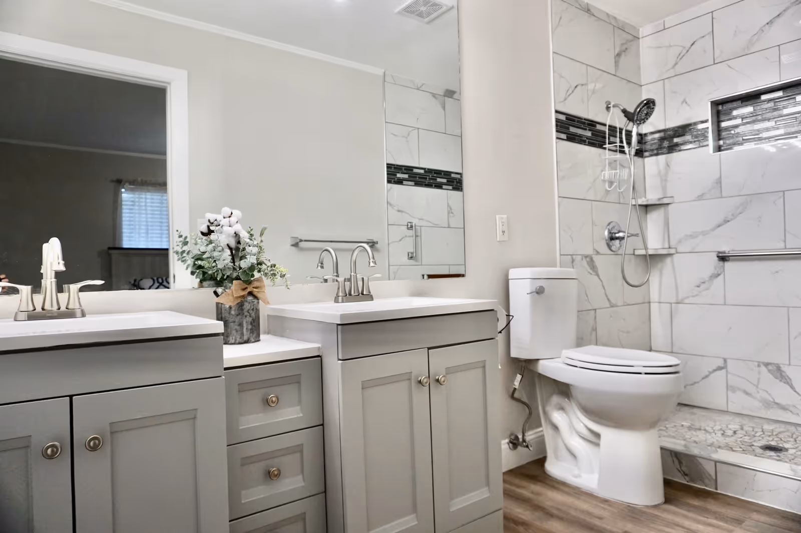 A modern bathroom featuring two gray vanities with white countertops and silver faucets, a decorative plant in a vase between the sinks, a toilet, and a walk-in shower with marble-like tiles and a built-in shelf. The shower has a handheld showerhead and a horizontal accent strip of dark tiles. The floor is wood-style, and a large mirror spans the wall above the vanities.