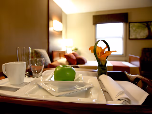 A close-up view of a tray with a green apple on a white plate, a white cup, two empty glasses, a rolled white napkin, and a small vase with orange flowers. In the background, there is a cozy living area with a sofa, cushions, a window with a brown valance, and a lit wall lamp.