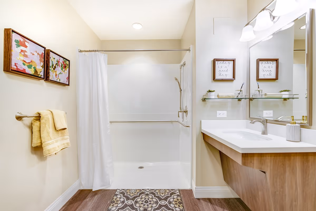 A clean and modern bathroom with a walk-in shower featuring a white curtain and grab bars. There is a wooden vanity with a white countertop and a sink, a large mirror above it, and a light fixture with three bulbs. Two framed abstract paintings hang on the wall above a towel rack with yellow towels. The floor has wood-like flooring with a patterned rug in front of the shower.