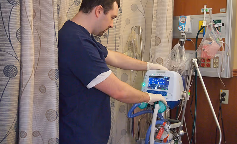 A healthcare worker in navy scrubs and gloves operates a medical ventilator machine in a clinical setting with privacy curtains and medical equipment visible in the background.