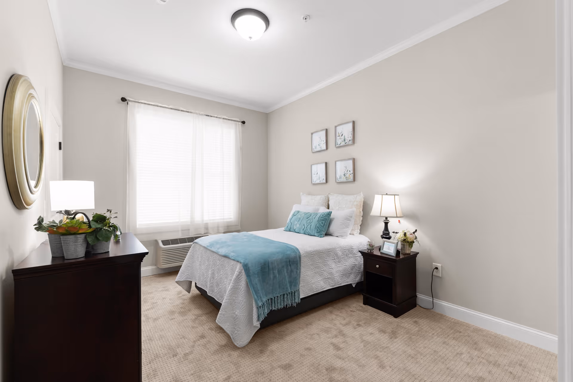 A neatly arranged bedroom with a bed covered in white bedding and a light blue throw blanket and pillow. There is a dark wooden nightstand with a lamp and small decorative items on the right side of the bed. On the left side of the room, there is a dark wooden dresser with a round mirror above it and a potted plant on top. The room has beige carpet, light-colored walls, and a window with sheer white curtains letting in natural light.