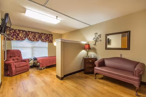 A cozy room with wooden flooring featuring a maroon armchair and a hospital-style bed near a window with floral curtains. A small partition separates the bed area from a sitting area with a maroon upholstered loveseat, a wooden nightstand with a lamp, and a wall-mounted mirror.