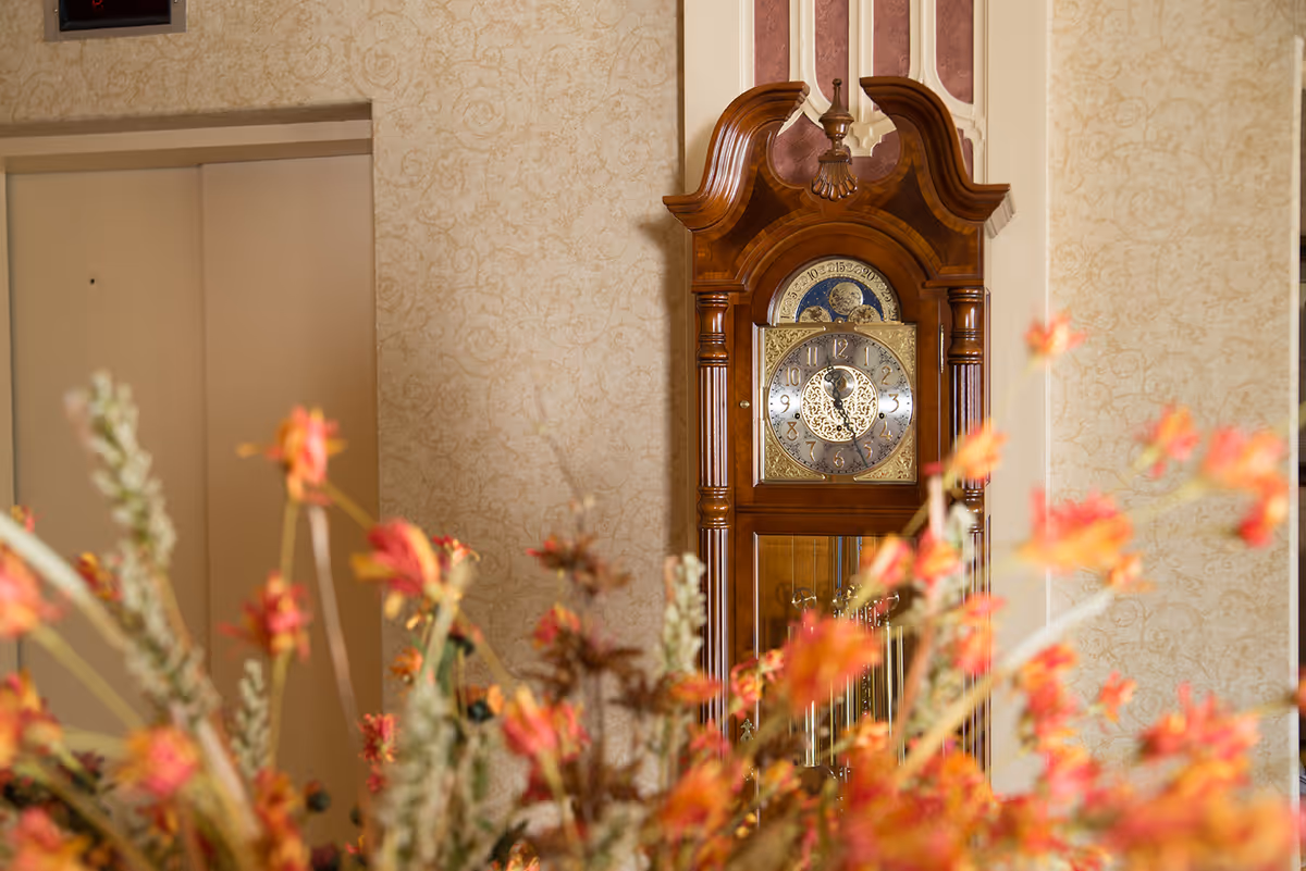 A wooden grandfather clock with an ornate design stands against a wallpapered wall with a floral pattern. In the foreground, there are blurred orange and red flowers. To the left of the clock, there is a closed elevator door.