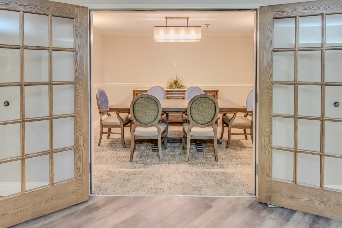 View through open wooden framed glass double doors into a dining room with a rectangular wooden table surrounded by six upholstered chairs. A modern rectangular light fixture hangs above the table, and a sideboard with a decorative plant is against the far wall. The room has light-colored walls and a patterned carpet.