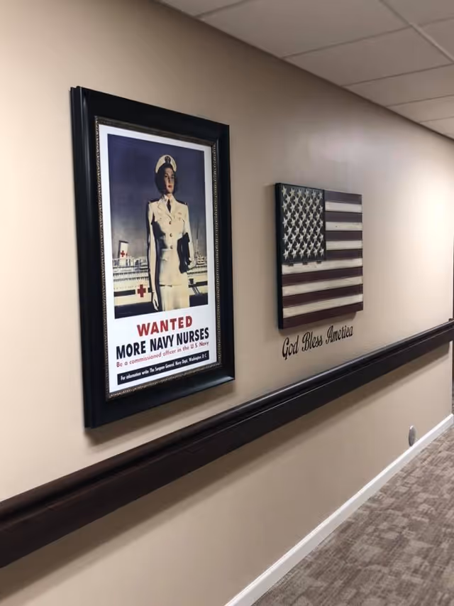 Hallway interior wall displaying a framed vintage Navy nurses recruitment poster and a wooden American flag with the words 'God Bless America'.