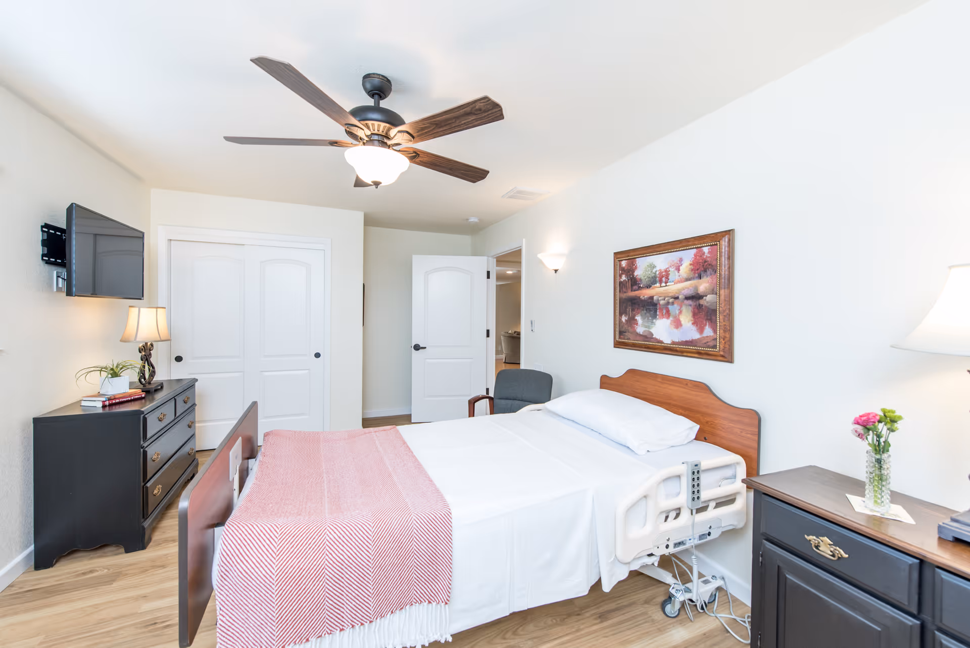 Bright private bedroom in a senior living facility with a medical adjustable bed, dresser, wall-mounted TV, ceiling fan, and framed artwork.
