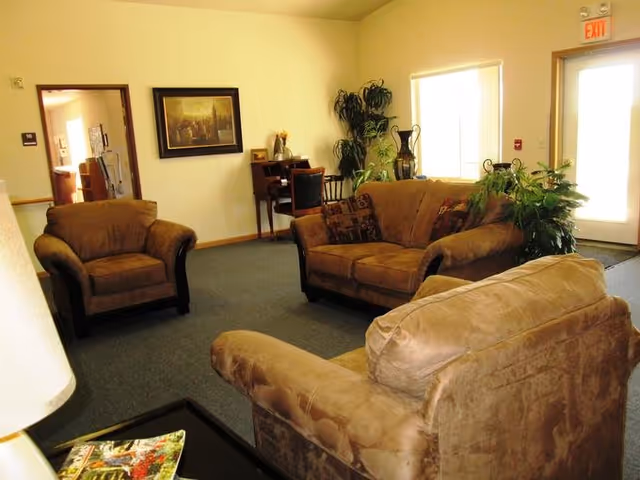A cozy living room area with three brown upholstered chairs and a loveseat arranged around a coffee table. There is a small desk with a chair against the far wall, a large plant near a window, and a framed picture hanging on the wall. An exit door with a window is visible on the right side.