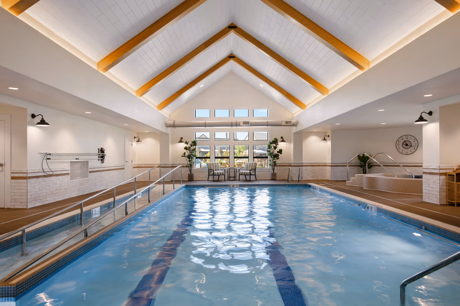 Indoor swimming pool with clear blue water, a ramp with handrails for accessibility, and a hot tub area to the right. The room has a high vaulted ceiling with wooden beams, large windows at the far end letting in natural light, and seating with two chairs and a small table near the windows. The walls are white with light-colored tile accents and there are several wall-mounted lights.