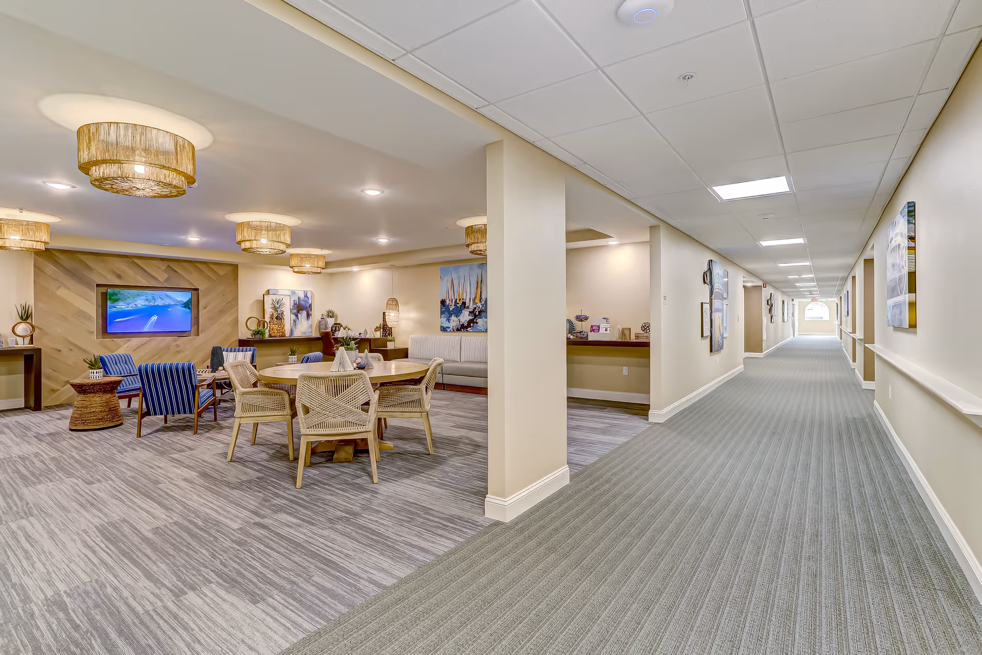 Bright communal lounge area with round tables, wicker and upholstered chairs, pendant lights, a TV on a wood-accent wall, and a long carpeted hallway leading away.