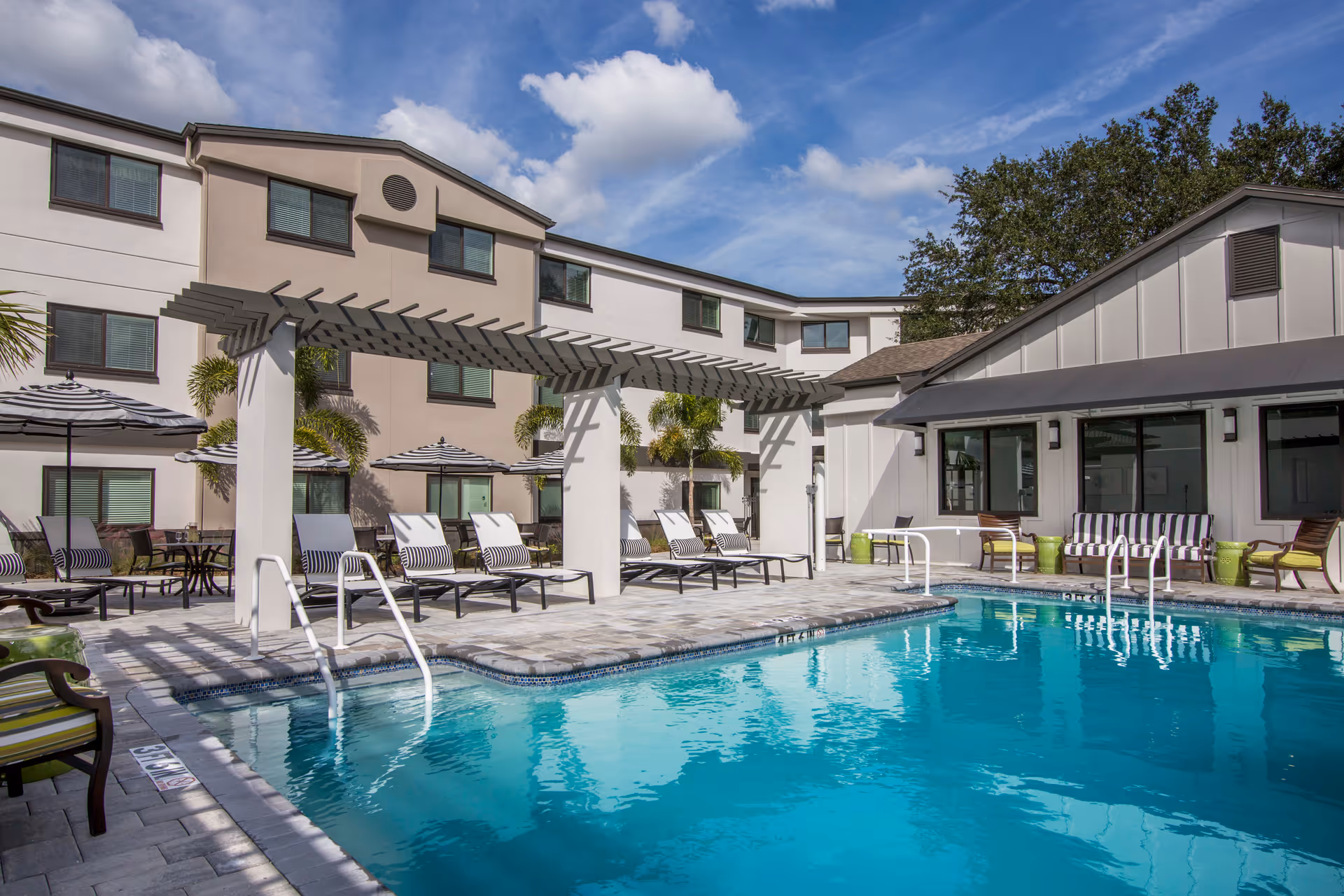 Outdoor swimming pool area at Renaissance North Tampa with lounge chairs, striped umbrellas, a pergola, and a building in the background under a partly cloudy sky.