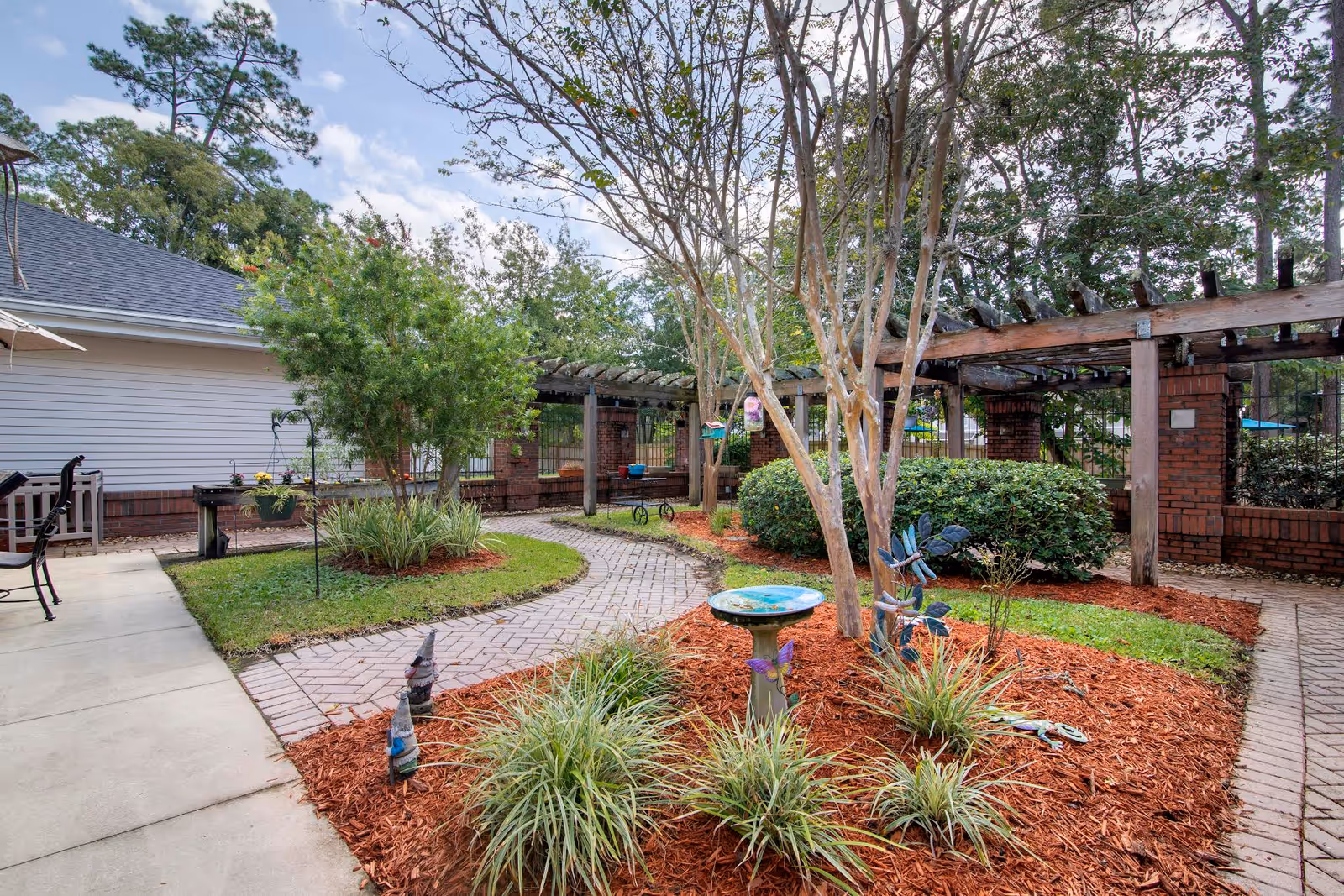 Outdoor courtyard with a brick walkway, planted beds, a birdbath, and a wooden pergola.