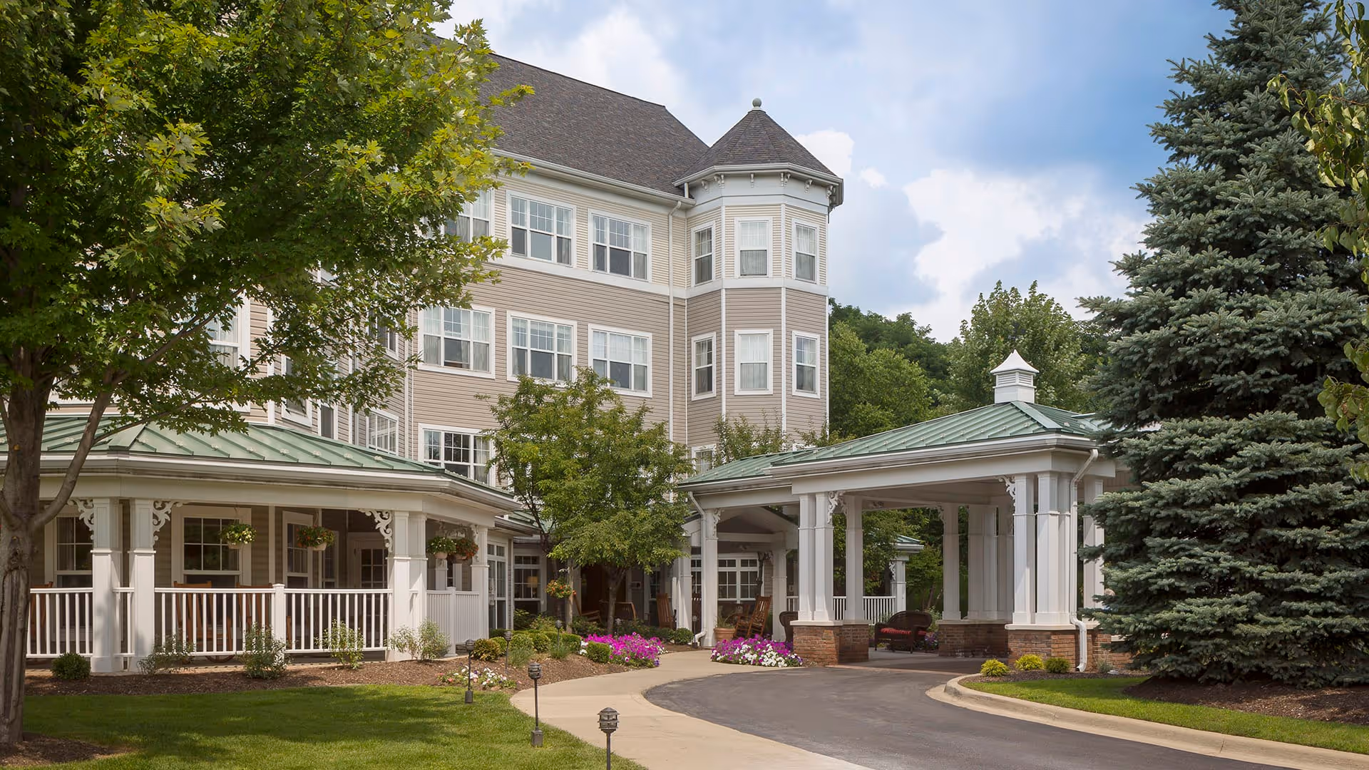 Exterior view of a senior living facility building with beige siding and white trim. The building has multiple windows and a turret feature. There is a covered driveway entrance with white columns and a green roof. Surrounding the building are well-maintained trees, shrubs, and flower beds under a partly cloudy sky.