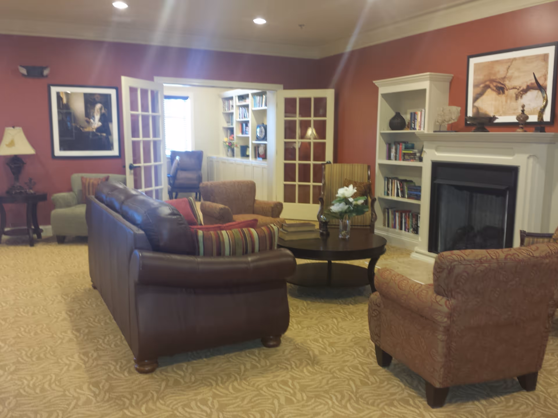 A cozy living room with a brown leather sofa, patterned armchairs, a round coffee table with a vase of flowers, built-in bookshelves, a fireplace, and French doors leading to a bright room with more seating and bookshelves.