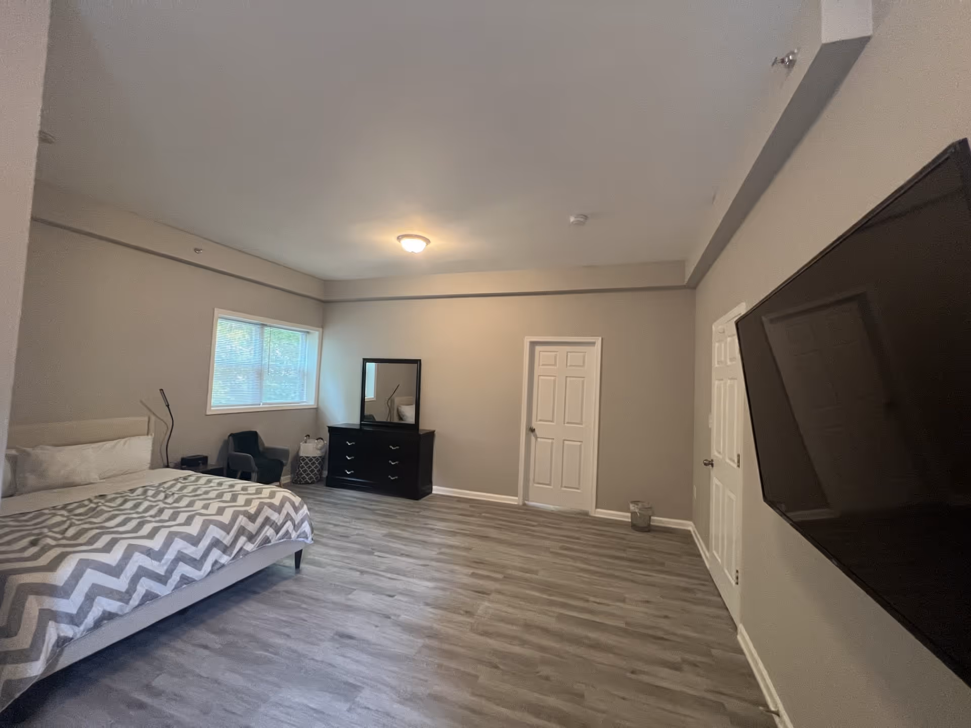 A spacious bedroom with a large bed covered in a gray and white chevron patterned blanket, a window with blinds, a black dresser with a mirror, a small chair, and a wall-mounted flat-screen TV. The room has light gray walls and wood-look flooring.
