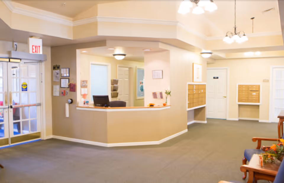 Reception area of a senior living facility with a front desk, computer, mailboxes on the walls, seating area with chairs and a table with flowers, beige walls, carpeted floor, and overhead lighting. Glass double doors with an exit sign are visible on the left.