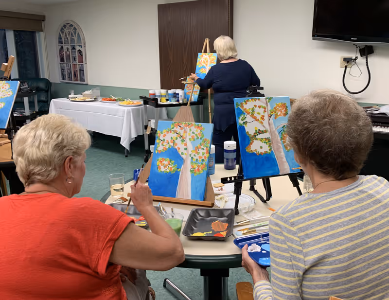 Three elderly women engaged in a painting activity in a room. Two women are seated at a table with easels, painting trees with colorful leaves on blue backgrounds. A third woman stands at an easel in the background, also painting a similar tree. The room has green carpet, a table with snacks covered by a white tablecloth, and a wall-mounted TV.