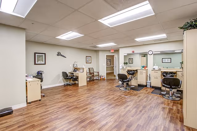 Interior view of a salon area in a senior living facility with wooden flooring, several salon chairs, mirrors, and workstations. The room is well-lit with ceiling lights and decorated with framed pictures on the walls.