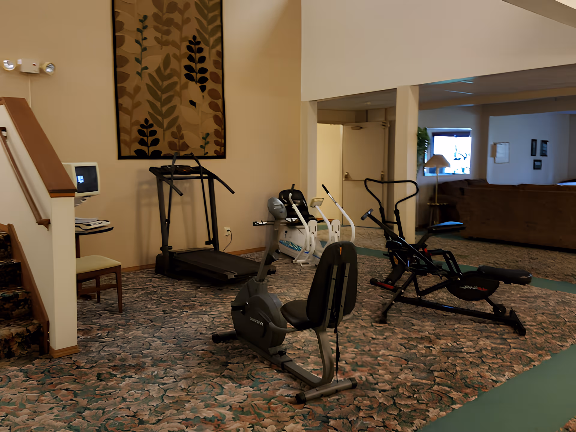 Interior fitness area with a treadmill, recumbent bike, and rowing machine in a retirement community lounge.