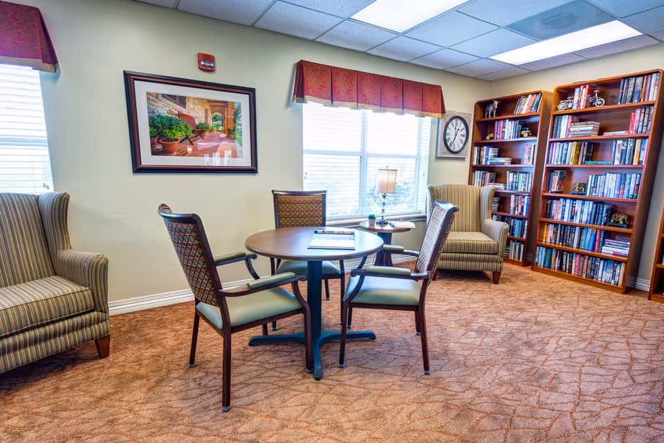 A cozy reading and sitting area in a senior living facility with a round wooden table surrounded by four chairs, two striped armchairs, a large window with red valances, a table lamp, a wall clock, framed artwork, and two wooden bookshelves filled with books and decorative items.