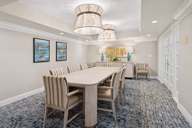 A well-lit dining room with a long rectangular table surrounded by eight striped upholstered chairs. The room features two large pendant lights hanging from the ceiling, a patterned blue carpet, two framed abstract paintings on the left wall, and a sideboard with decorative lamps and a flower arrangement at the far end. French doors are visible on the right side of the room.