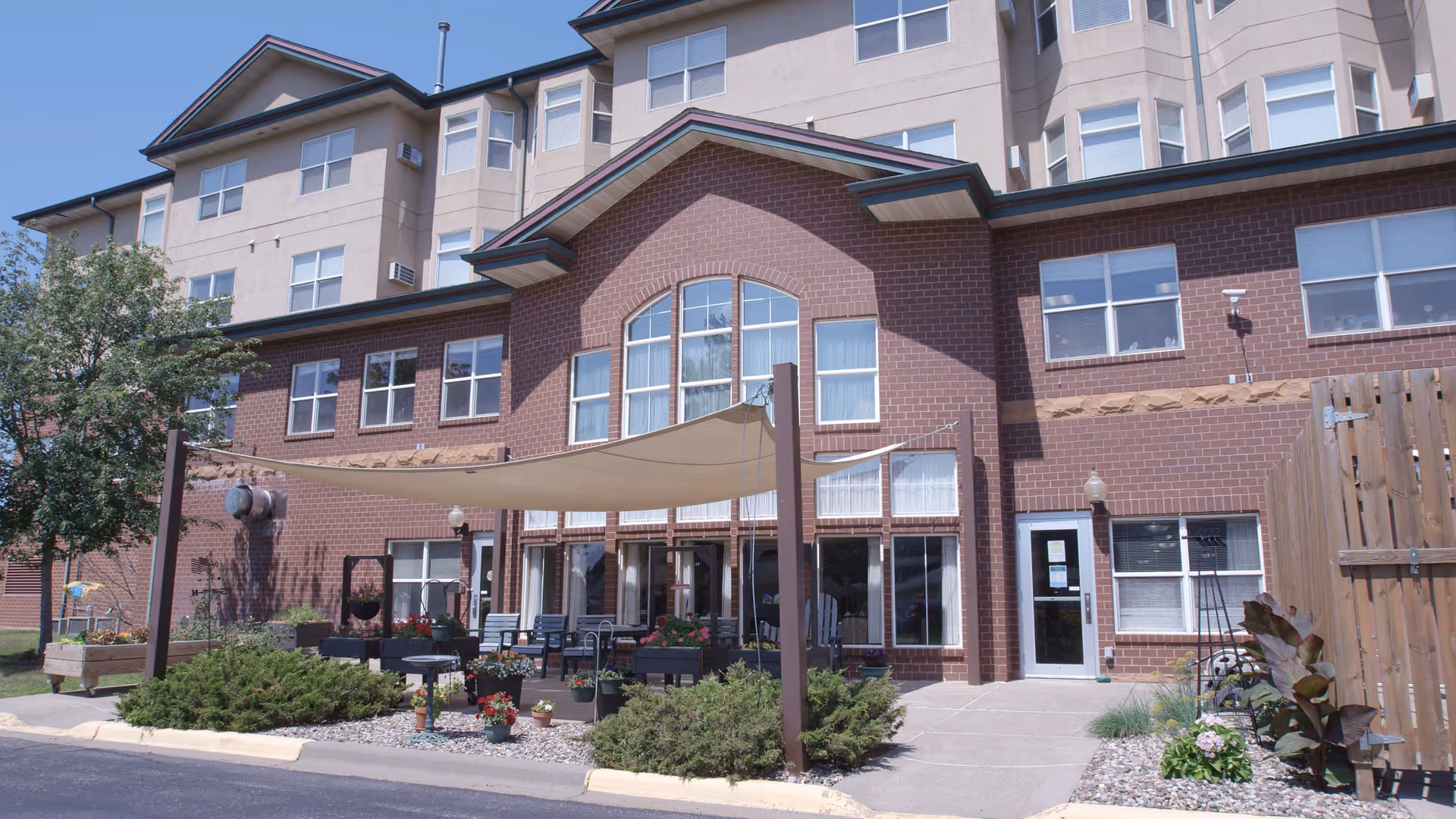 Front exterior of a multi-story brick senior living building with a shaded patio area, outdoor seating, planters, and landscaping.