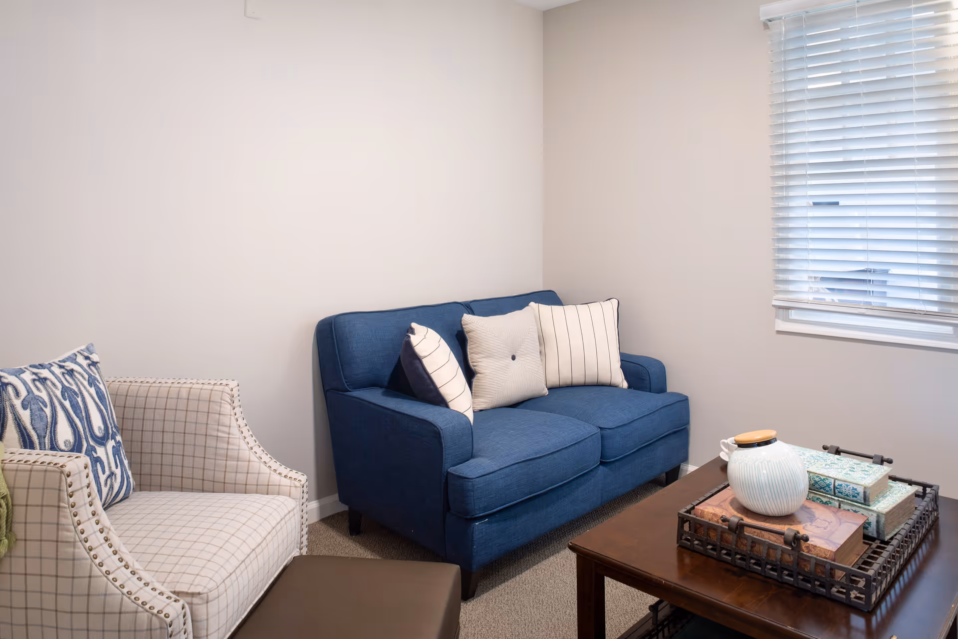 A small living room with a blue loveseat adorned with three decorative pillows, a beige armchair with a blue patterned pillow, a dark brown ottoman, and a wooden coffee table holding a tray with a white ceramic pitcher and decorative boxes. A window with white blinds is on the right wall.