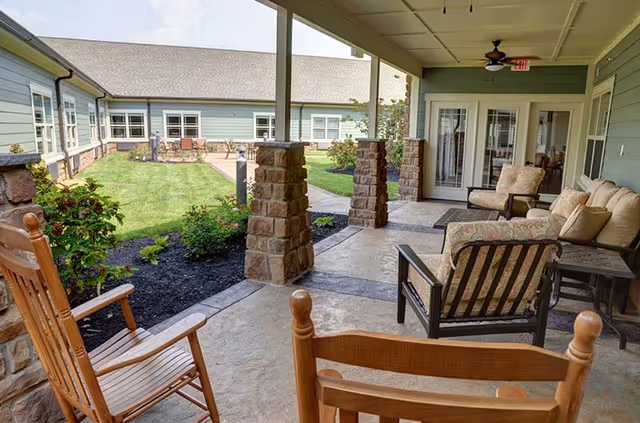 Covered outdoor patio area at Clover Hill Senior Living with wooden rocking chairs and cushioned armchairs arranged for seating. Stone pillars support the roof, and a well-maintained lawn and garden are visible beyond the patio. The building exterior is light green with multiple windows and a glass door leading inside.