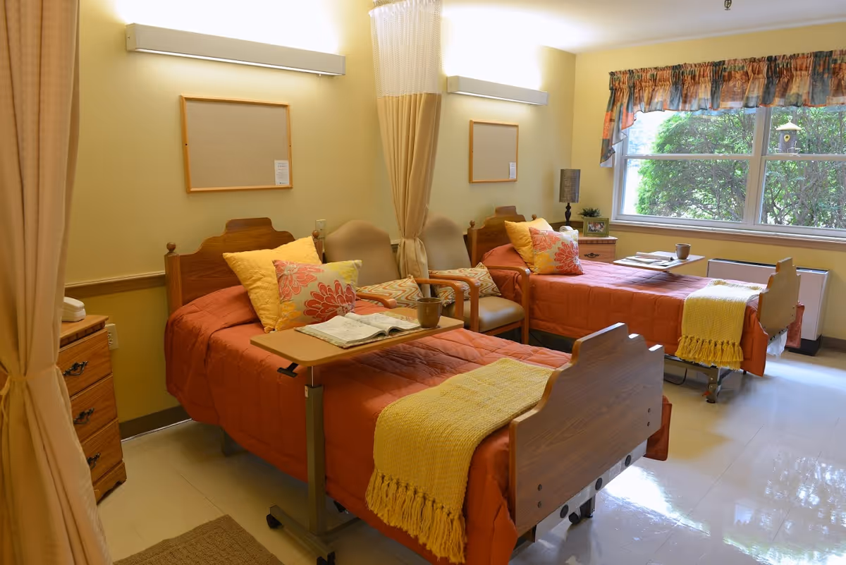A bright room with two hospital-style beds, each with orange bedspreads, yellow throws, and decorative pillows. Between the beds are two beige chairs and a small table with a lamp and picture frame. A large window with floral curtains lets in natural light and shows greenery outside. Privacy curtains hang from the ceiling around the beds.