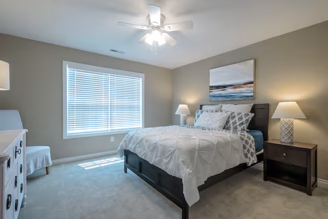 A bright bedroom with a large window covered by white blinds letting in natural light. The room features a bed with white and patterned bedding, two bedside tables each with a lamp, a white dresser, and a white upholstered chair. The walls are painted beige and there is a ceiling fan with a light fixture.