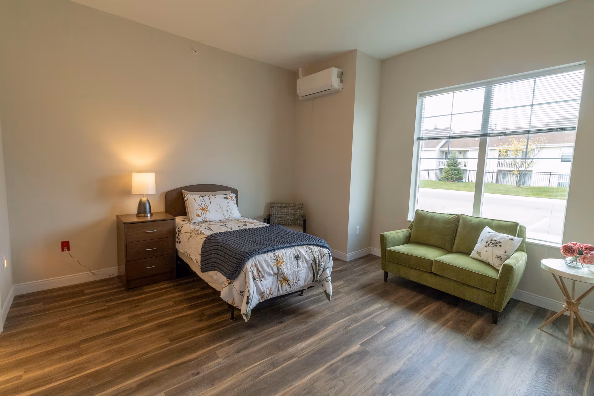 A bright and clean bedroom with a single bed featuring floral bedding and a gray throw blanket. Next to the bed is a wooden nightstand with a lit lamp. A green loveseat with a decorative pillow is placed near a large window with blinds, allowing natural light to fill the room. The floor is covered with wood-style flooring, and there is a small round side table with a vase of pink flowers beside the loveseat. An air conditioning unit is mounted on the wall above the chair in the corner.