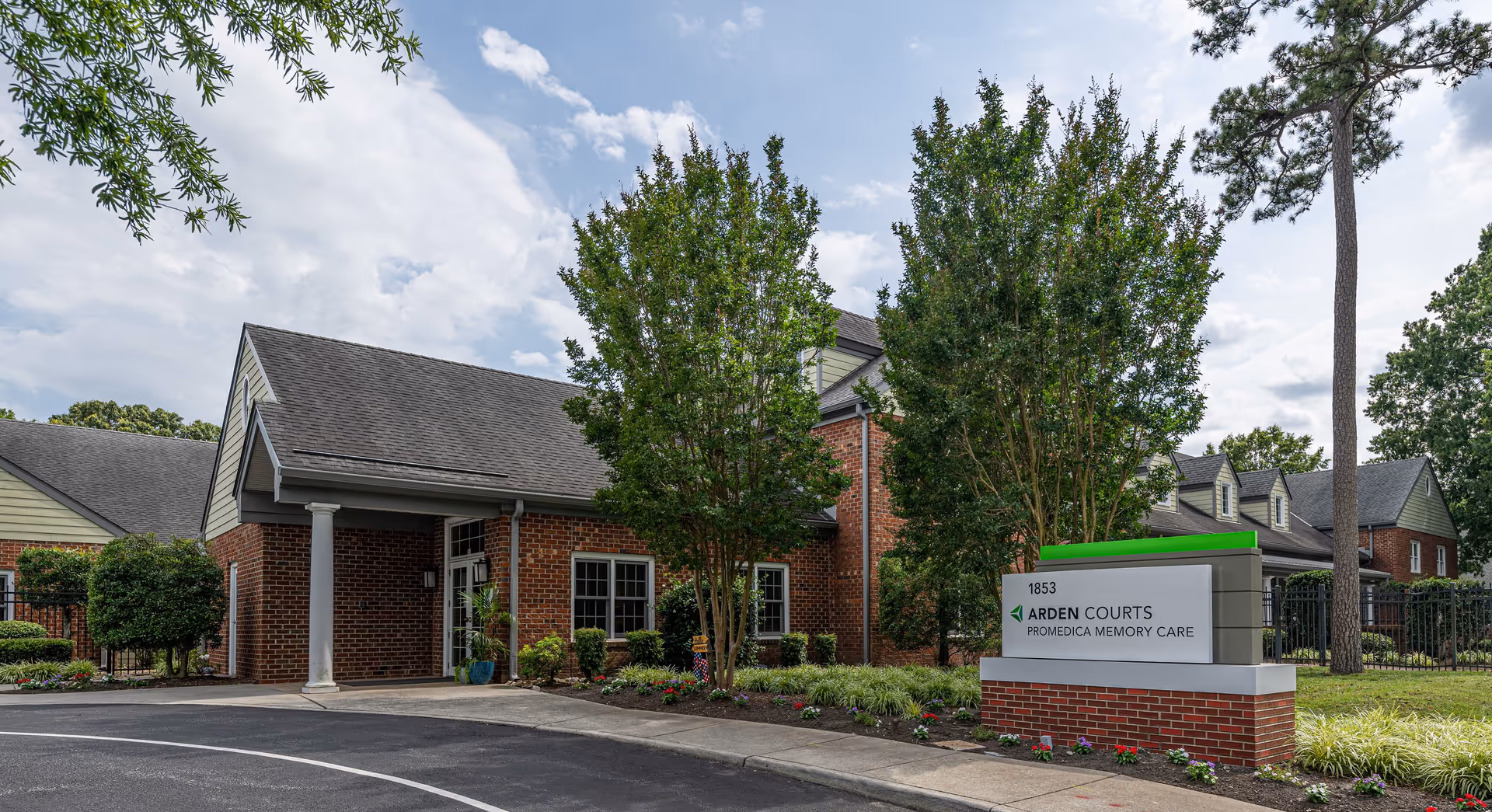 Exterior view of Arden Courts - ProMedica Memory Care Community building in Virginia Beach, showing a brick facade with a covered entrance, surrounded by trees and landscaping under a partly cloudy sky.