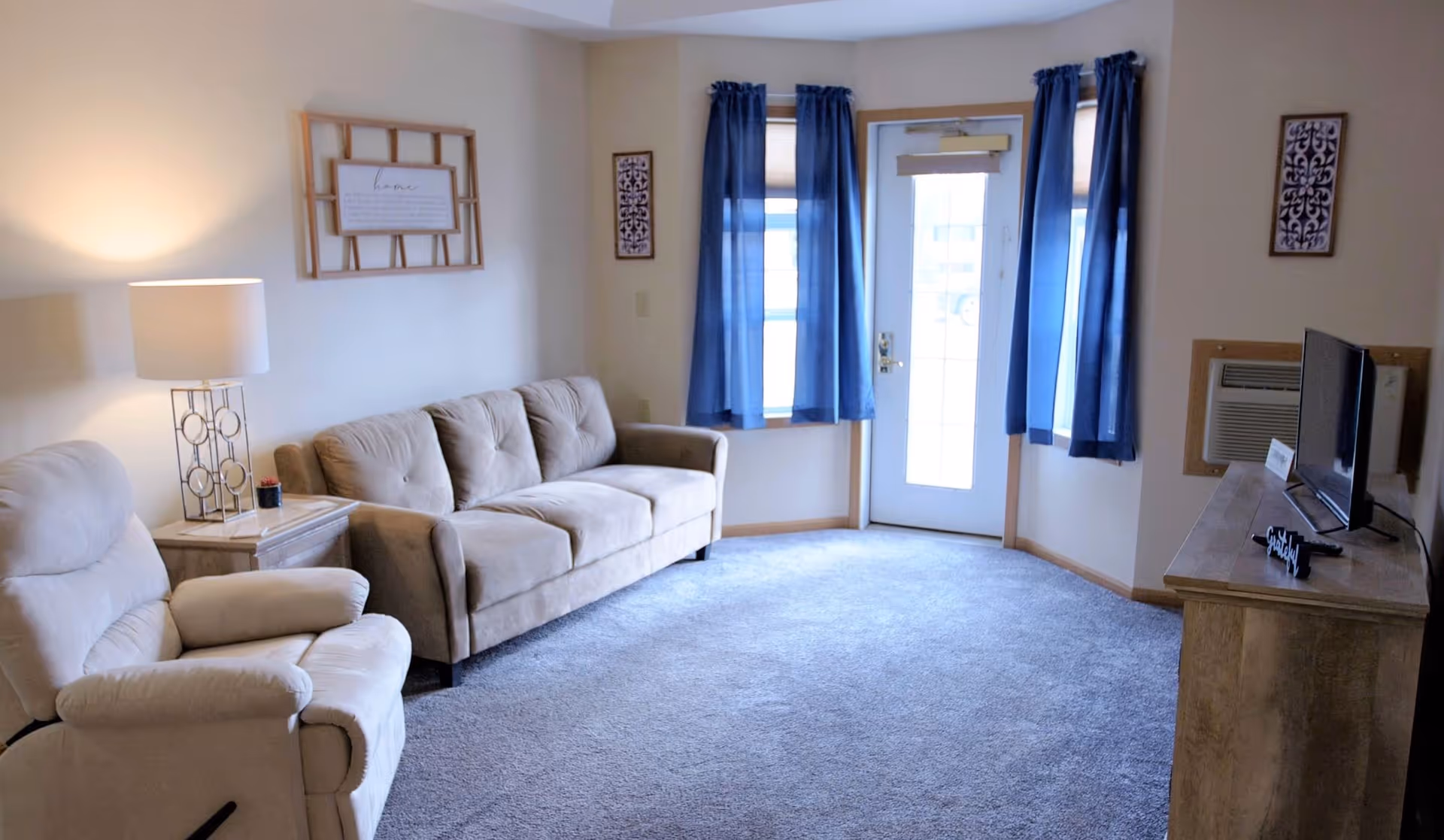 Cozy living room with a beige sofa and recliner, side table and lamp, TV on a wooden console, blue curtains and a glass exterior door.