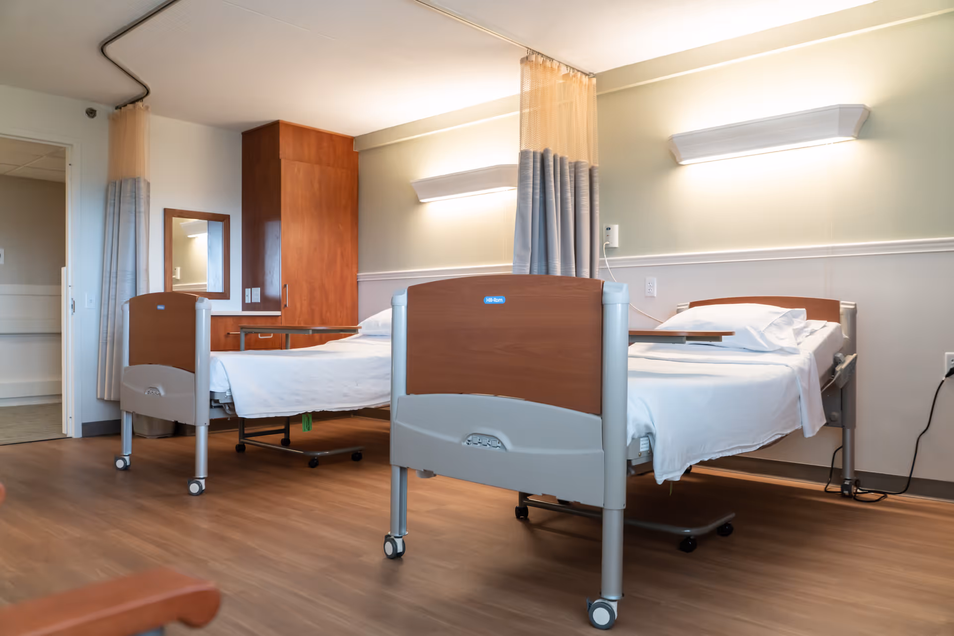 A clean and well-lit room with two hospital-style beds, each with white sheets and pillows. The beds have wooden headboards and footboards with metal frames on wheels. There is a wooden cabinet and a mirror on the far wall, and privacy curtains hanging from the ceiling between the beds. The floor is wood laminate, and the walls are painted light green and white with wall-mounted lights above each bed.