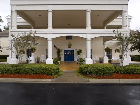 Front exterior view of a building with white columns and a covered porch area featuring several white rocking chairs and hanging plants. There are shrubs and small trees in front of the porch, and a blue double door entrance in the center.