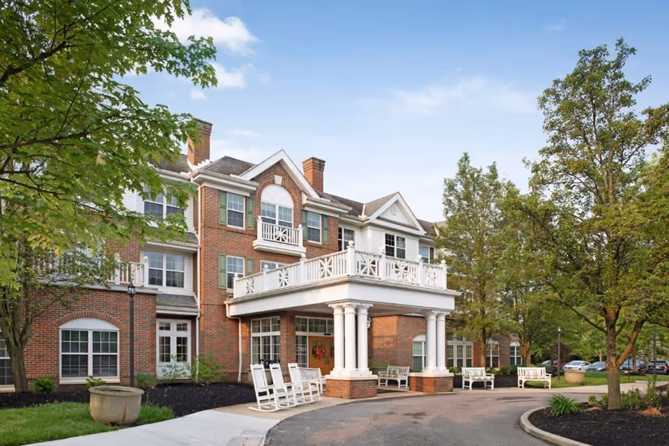 Exterior view of a senior living facility building with red brick and white trim, featuring a covered entrance supported by white columns. Several white rocking chairs and benches are placed near the entrance. Trees and greenery surround the building under a partly cloudy sky.