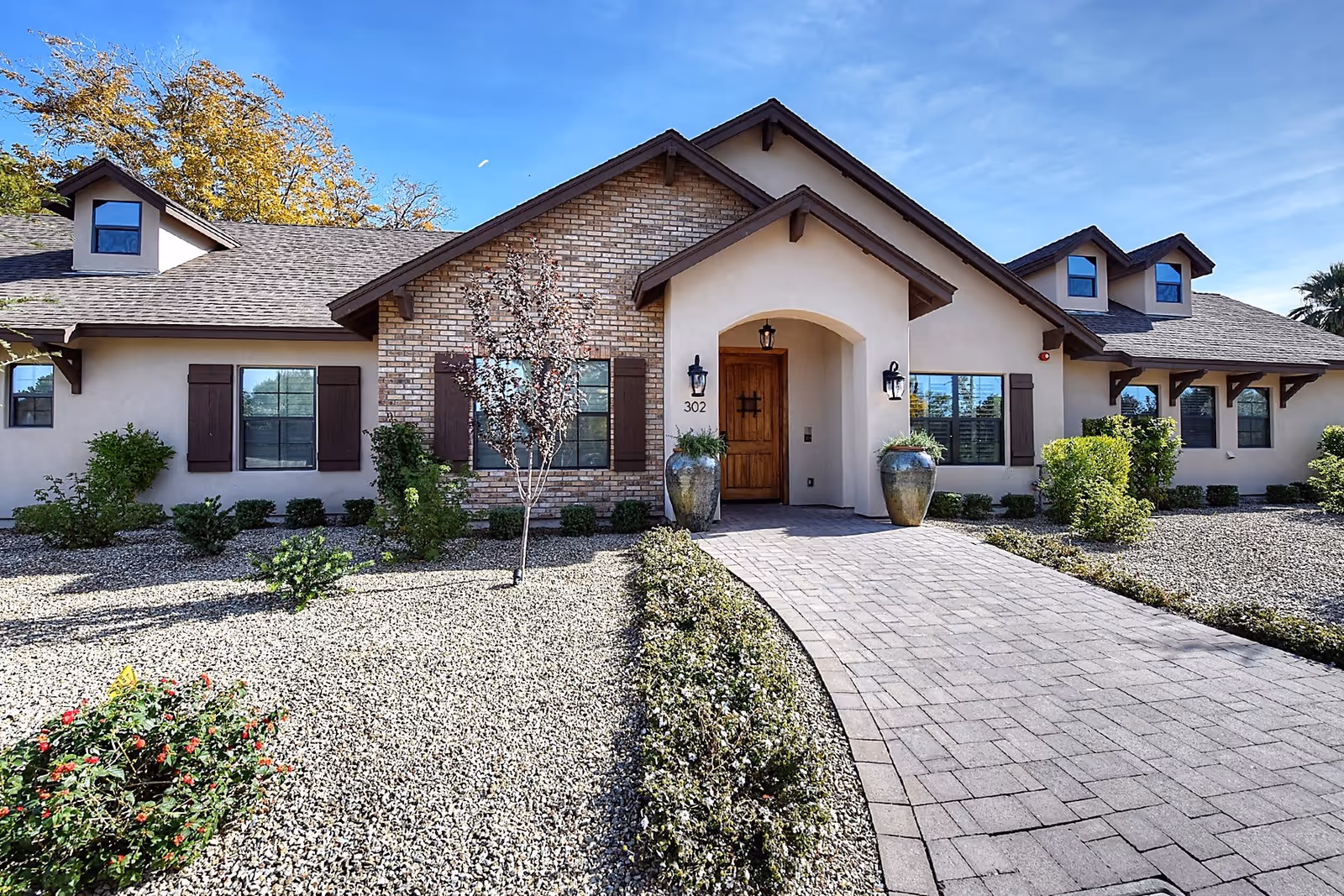 Front exterior view of a single-story building with beige walls, brown roof shingles, and multiple windows with brown shutters. A paved walkway leads to a wooden front door under an arched entryway, flanked by two large decorative pots with plants. The surrounding landscape features gravel ground cover, small bushes, and a few small trees under a clear blue sky.