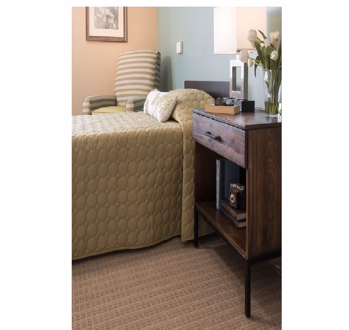A cozy bedroom corner featuring a bed with a quilted beige bedspread and decorative pillows, a wooden nightstand with a drawer and open shelf holding books and a vintage camera, a modern table lamp, a vase with white tulips, and a striped armchair against a two-tone wall with a framed floral artwork.