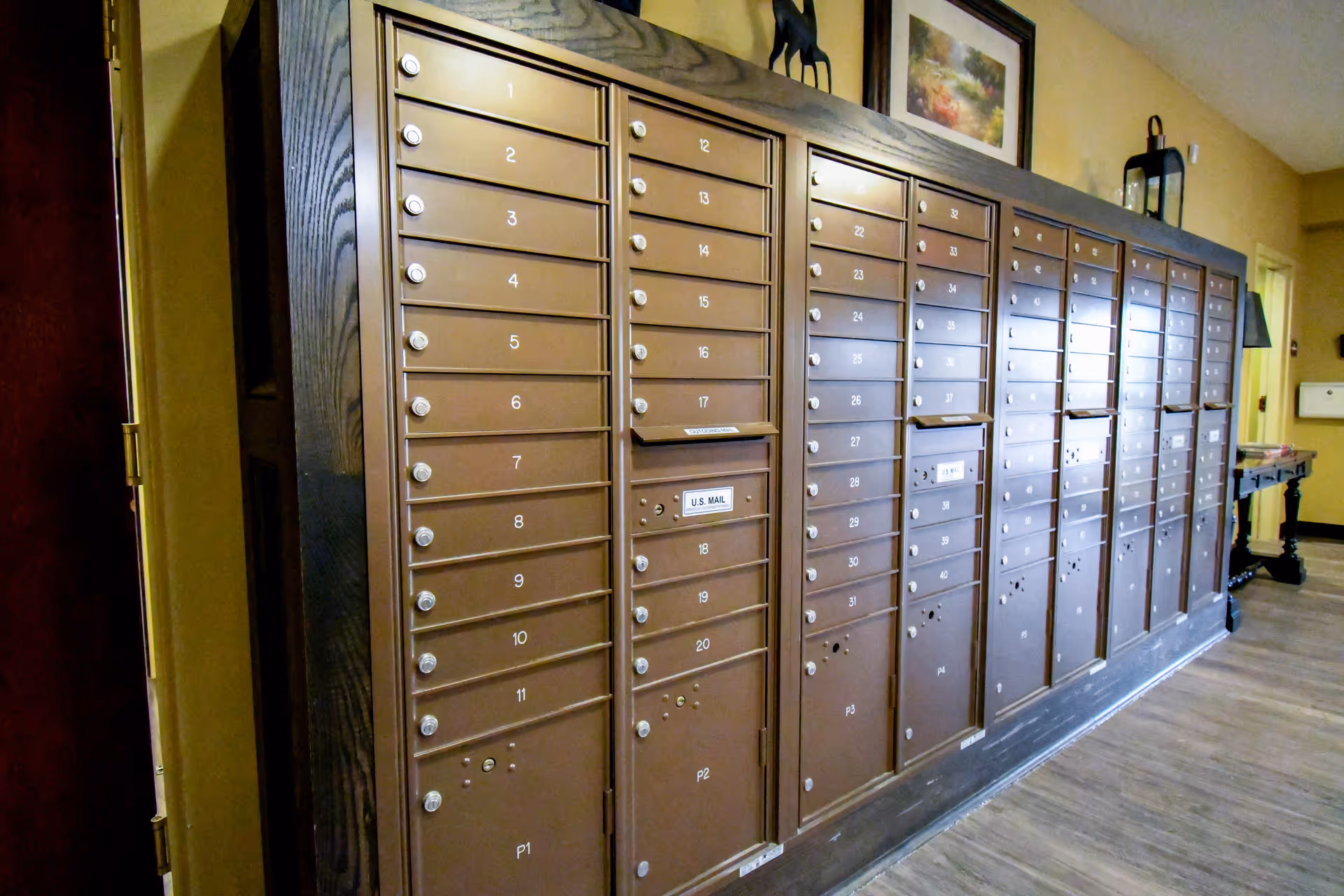 A row of brown metal mailboxes with numbered compartments inside a hallway with wooden flooring and beige walls. There is a framed picture and a lantern on top of the mailbox unit.