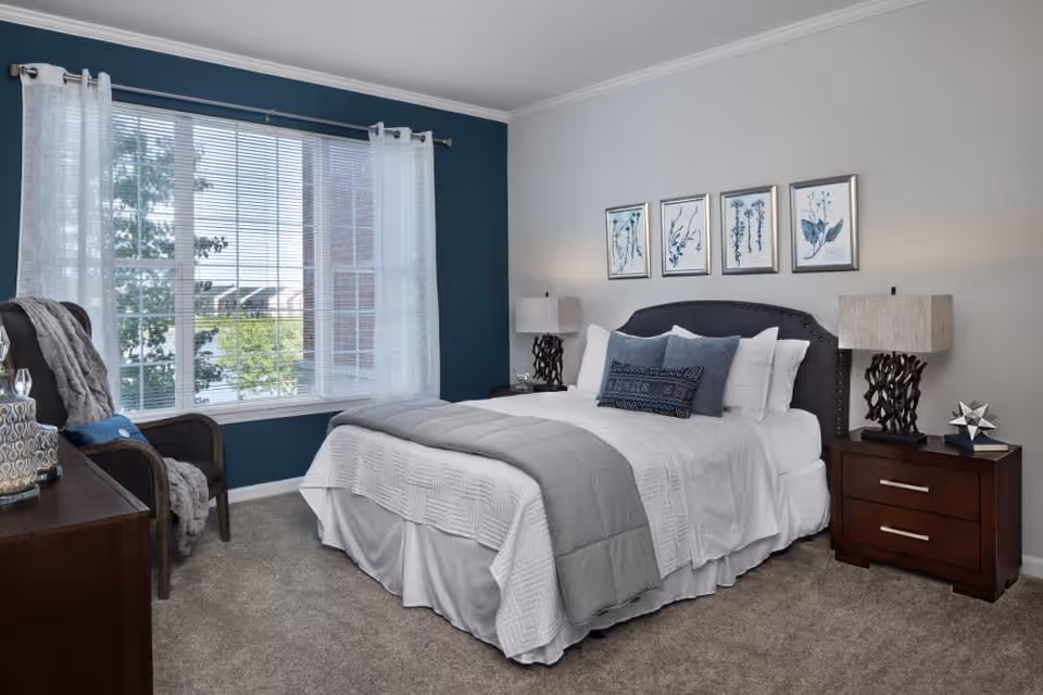 A neatly made bed with white and gray bedding in a bedroom. The bed has a dark upholstered headboard and is flanked by two wooden nightstands with matching lamps. Above the bed are four framed botanical prints. A large window with white curtains and blinds lets in natural light. There is a dark armchair with a gray throw blanket and a wooden dresser with decorative items on the left side of the room. The walls are painted in a combination of blue and light gray.