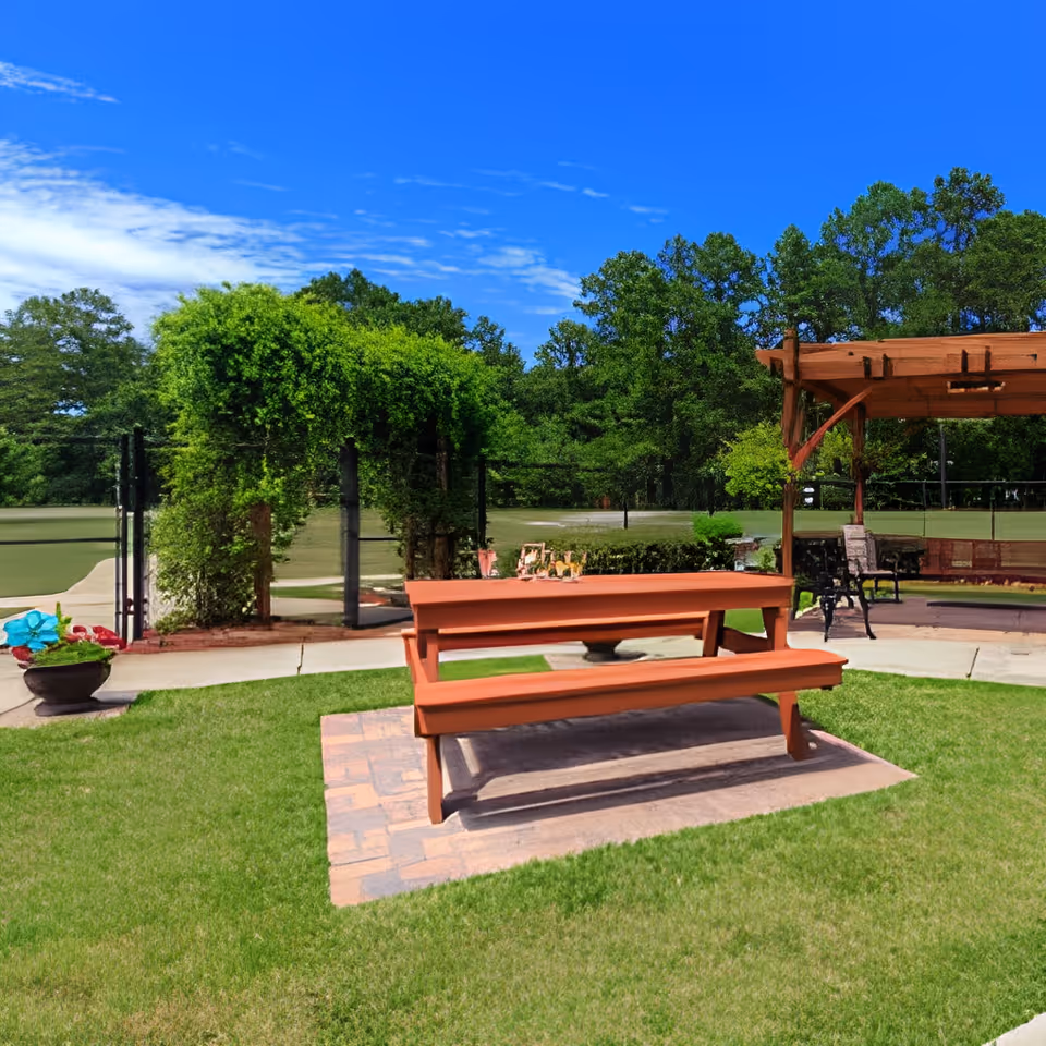 Outdoor courtyard with a wooden picnic table on a grassy lawn, a pergola and trees in the background.