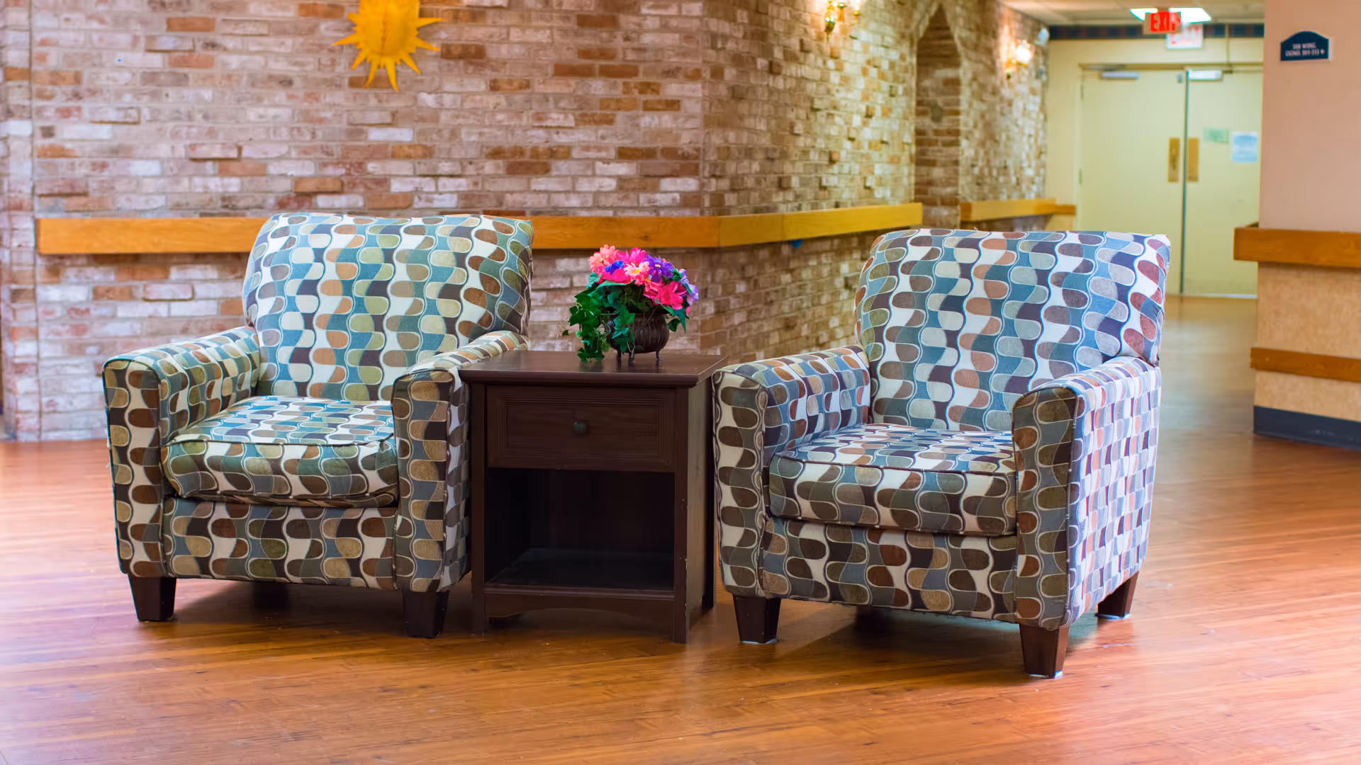 Two patterned armchairs with a retro geometric design are placed on either side of a small dark wooden side table with a drawer and an open shelf. A small flower arrangement with pink and purple flowers sits on the table. The setting is indoors with a wooden floor, brick wall, and hallway in the background.