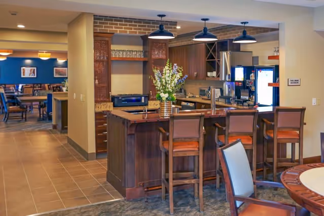 Interior view of a senior living facility's kitchen and dining area featuring a wooden kitchen island with four high chairs, a vase with flowers on the island, modern kitchen appliances, and a dining area with tables and chairs in the background.